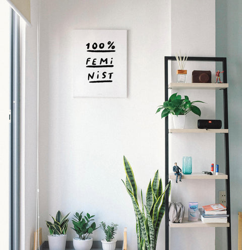 Feminist poster for women painted with black ink and hand lettered typography saying "100% feminist" and hanging on the wall in a feminine, modern and bright room.