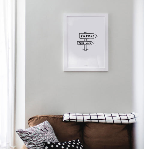 Black and white comic style drawing featuring a street sign with an arrow pointing to the future. It says "Future, this way". An inspirational wall art hanging above the sofa in a living room.