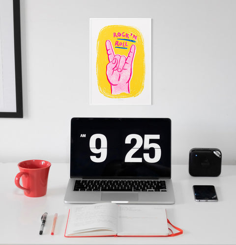 Pink and yellow retro music wall art print featuring an illustration of a hand with two fingers up and hand lettered word Rock'n roll. This graphic illustration art print is hanging above a modern minimalist desk in a home office space.
