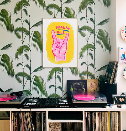 Music poster in eclectic living room hanging over desk with vinyl record player. The music wall art shows a hot pink hand and hand painted typography saying "rock and roll" on yellow background. Cool rock music wall art prints for living room.