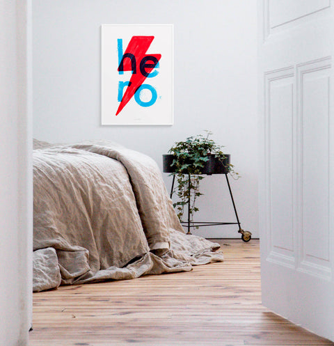 Bright and bold typography poster featuring hand lettered font saying Hero and a red lightning bolt. The minimalist print is hanging on the wall above bed in a modern bedroom.
