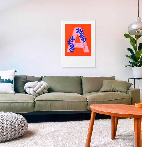 Initial letter A poster print hanging above sofa in nordic living room. Big letter A in pink with electric blue botanical details.