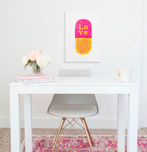Pink and yellow illustration wall art print featuring a cute love pill and the hand drawn word Love. This graphic art print is hanging above a minimalist desk in a white and pink home office space.
