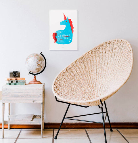 Inspirational wall art featuring an illustration of a cute unicorn and hand drawn letters saying I believe in  you. This bright and colorful illustration art print is hanging in a modern room.