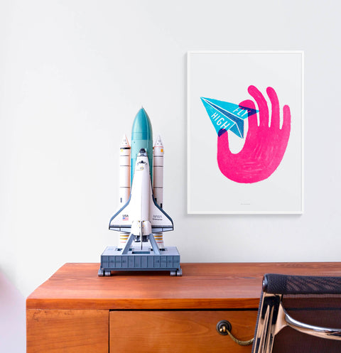 Inspirational wall art print featuring an illustration of a pink hand holding a blue paper plane with the words Fly high. The poster is hanging above a desk in an work from home office.