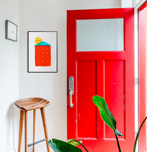 Graphic illustration wall art print featuring a simple red house and the word Home. This bright and bold graphic wall art is hanging in a bright entryway with a red door.