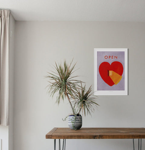 Open heart wall art is a bold illustration art print featuring a big red heart with a little open door and a golden ray of light coming out of it. A big red heart wall art hanging on a modern living room wall.