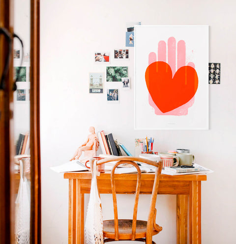 Red heart and pink hand illustration art print. This bold and bright graphic wall art is hanging above a wooden desk in a contemporary artsy office space.