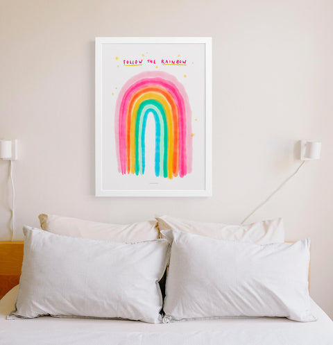 Watercolor rainbow poster with hand drawn phrase saying Follow the rainbow. This bright and colorful illustration print is hanging above the bed in a minimalist cosy bedroom.