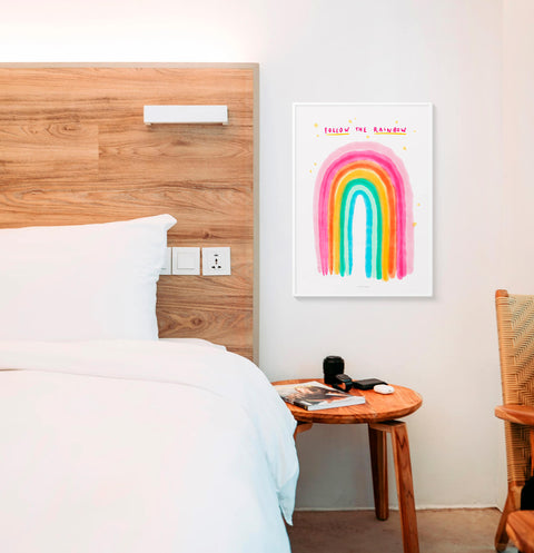 Colorful watercolor rainbow art print with hand drawn phrase saying Follow the rainbow. This cute illustration print is hanging above the bed in a modern bedroom.