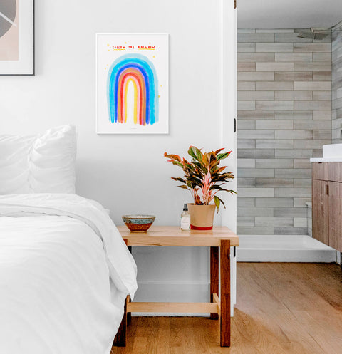Watercolor rainbow wall art print with hand lettered quote saying Follow the rainbow. This rainbow illustration print is hanging above a white bed in a modern bedroom.